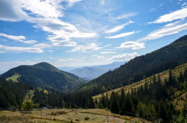 Ormanla kaplı yamaçlar, tepeler arasında bir vadi ve parçalı bulutlu mavi bir gökyüzü içeren güzel bir dağ manzarası dingin ve ferahlatıcı bir ortam yaratıyor. Karpat Dağları, Ukrayna 