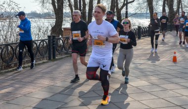 Bir maraton etkinliğinde birlikte koşan bir grup insan ağaçlarla çevrili asfalt bir yolda, spor yapmayı, dayanıklılığı ve aktif bir yaşam tarzını teşvik ediyorlar.