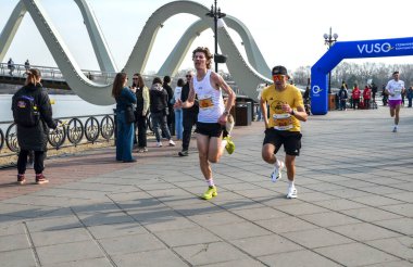 Koşucular, izleyiciler ve şehir zeminiyle çevrili, belirgin yapılandırılmış bir köprünün yakınındaki bir maraton etkinliğinde yarışıyorlar.