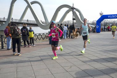 Koşucular, izleyiciler ve şehir zeminiyle çevrili, belirgin yapılandırılmış bir köprünün yakınındaki bir maraton etkinliğinde yarışıyorlar.