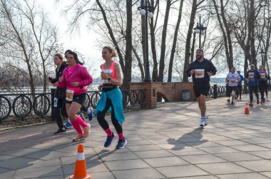 Bir maraton etkinliğinde birlikte koşan bir grup insan ağaçlarla çevrili asfalt bir yolda, spor yapmayı, dayanıklılığı ve aktif bir yaşam tarzını teşvik ediyorlar.