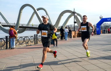 Koşucular, izleyiciler ve şehir zeminiyle çevrili, belirgin yapılandırılmış bir köprünün yakınındaki bir maraton etkinliğinde yarışıyorlar.