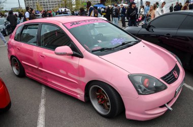 Modifiye edilmiş, pembe sakızlı Honda Civic hatchback park halinde, pazardan sonra gelen tekerlekler, indirilmiş bir duruş ve ön camında Japonca bir yazı pankartı var.