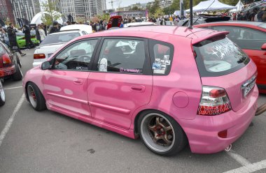 Modifiye edilmiş, pembe sakızlı Honda Civic hatchback park halinde, pazardan sonra gelen tekerlekler, indirilmiş bir duruş ve ön camında Japonca bir yazı pankartı var.