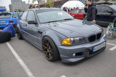 Bir mat gri BMW E46 sedan, geniş gövde kiti ile ağır modifiye, düşük süspansiyon ve bronz çok dilli tekerlekler, açık havada park edilir