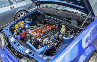 Canlı mavi bir Ford Sierra arabasının kaputu, farklı bir turuncu motor kapağı ve çok sayıda hortum ve bileşen içeren motor bölümünün detaylı bir görüntüsünü ortaya çıkarır.