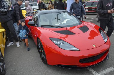 Siyah çatılı kırmızı Lotus Evora açık hava araba etkinliğinde sergileniyor. Pürüzsüz, aerodinamik tasarımı ve dikkat çekici arka spoiler spor arabasının cazibesini vurguluyor.