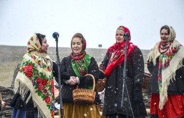 Geleneksel giysiler içinde bir grup kadın dışarıda, karlı bir gökyüzünün altında, Shrovetide kutlamaları sırasında toplumu ve kültürel gelenekleri sembolize ederek neşe içinde şarkı söylüyor.