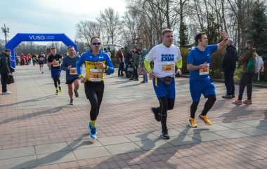 Bir açık hava etkinliği sırasında maraton katılımcıları asfalt bir patika boyunca aktif olarak koşuyorlar. Seyirciler onları gözlemler ve neşelendirir, canlı bir atmosfer yaratırlar.