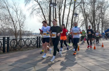 Açık bir günde kaldırımlı park yolunda koşan bir grup insan etkinlik, spor, sağlık ve açık hava enerjisini taşır.