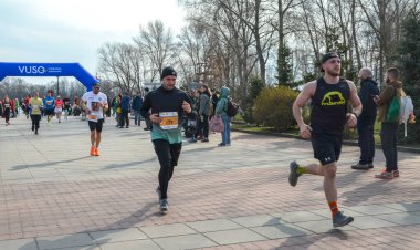 Bir açık hava etkinliği sırasında maraton katılımcıları asfalt bir patika boyunca aktif olarak koşuyorlar. Seyirciler onları gözlemler ve neşelendirir, canlı bir atmosfer yaratırlar.