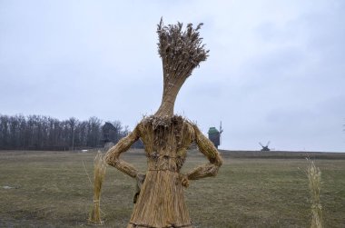 Geleneksel Ukrayna saman heykeli (Didukh), Shrovetide kutlamaları sırasında uzak arkaplanda geleneksel yel değirmenleri ve çıplak ağaçlarla bulutlu bir gökyüzünün altında uçsuz bucaksız, kuru bir alanda duruyor.