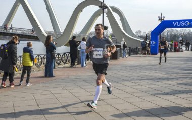 Nehir kenarındaki bir gezinti alanında düzenlenen bir maraton etkinliğine katılanlar, manzaralı bir arkaplanda seyirciler tarafından tezahürat yaparak aralarında çarpıcı bir modern köprü de yer aldı.