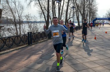 Gri uzun kollu tişörtlü ve siyah şortlu bir erkek koşucu bir yarış sırasında gülümsüyor ve baş parmağını kaldırıyor. Arka planda diğer koşucular, bir nehir ve çıplak ağaçlar var.