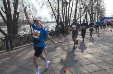 Mavi tişörtlü ve siyah şortlu bir bayan koşucu yarış sırasında kameraya el sallıyor. Arkasında bir nehir ve ağaçla diğer koşucular görülebiliyor.