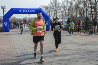 Koşucular organize maraton etkinliği sırasında kaldırımlı yolda birlikte koşuyorlar. Sahneye çeşitli yaşlardan katılımcılar, ağaçlar ve ilkbahar atmosferi katılıyor.