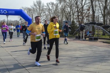 Koşucular organize maraton etkinliği sırasında kaldırımlı yolda birlikte koşuyorlar. Sahneye çeşitli yaşlardan katılımcılar, ağaçlar ve ilkbahar atmosferi katılıyor.