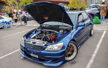 Lacivert Lexus IS, kaputu açık, ayrıntılı motoru açık, açık bir araba etkinliğinde sergileniyor. Piyasa sonrası tekerlekleri ve düşük bir duruşu var.