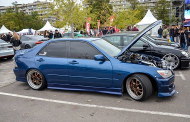 Lacivert Lexus IS, kaputu açık, ayrıntılı motoru açık, açık bir araba etkinliğinde sergileniyor. Piyasa sonrası tekerlekleri ve düşük bir duruşu var.