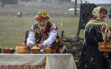 Geleneksel Ukrayna halk kıyafetleri içinde, renkli bir başlıkla, açık havada bir masaya kil kaplar koyuyor kar yağarken