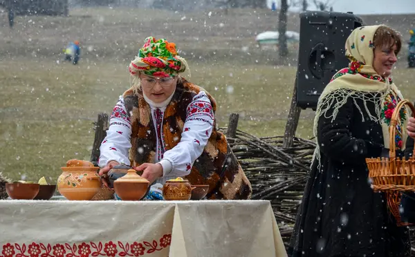 Geleneksel Ukrayna halk kıyafetleri içinde, renkli bir başlıkla, açık havada bir masaya kil kaplar koyuyor kar yağarken
