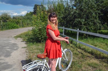 Beyaz bir bisikletin yanında duran neşeli bir kadın, kırmızı benekli bir elbise giymiş, güneşli bir günde yemyeşil ve ahşap çitli sakin bir kırsal bölgede.