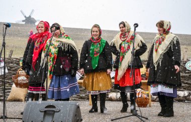 Karlı bir havada, geleneksel giysiler içinde açık havada şarkı söyleyen bir grup kadın. Sahne, şenlikli bir kültürel atmosfer yakalıyor. 