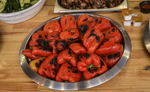Taze ızgara kırmızı biberler güzel kavrulmuş ve metal tabaklar üzerine organize edilmiş. Mantar ve salatalıklar da dahil. Canlı ve iştah açıcı yemek sunumu yapıyor.
