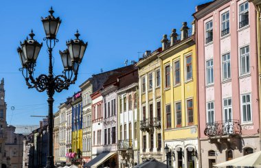 Market Meydanı 'nda açık mavi gökyüzünün altında çeşitli renklerde tarihi binalar var. Detaylı, süslü bir sokak lambası ön planda belirgin şekilde duruyor. Lviv, Ukrayna