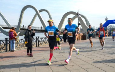 Rekabetçi bir maraton koşucuları arka planda belirgin bir köprü bulunan manzaralı bir patika boyunca ilerliyorlar.