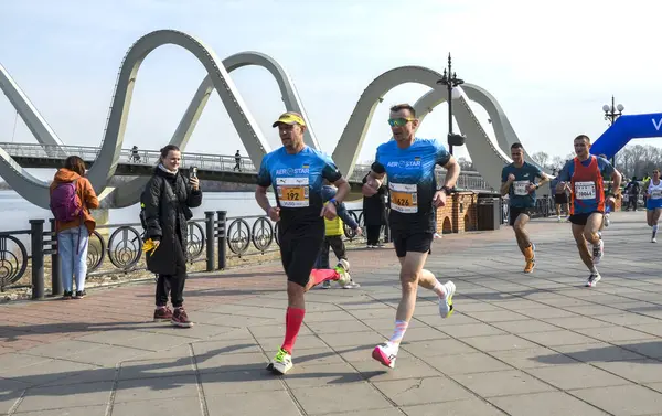Rekabetçi bir maraton koşucuları arka planda belirgin bir köprü bulunan manzaralı bir patika boyunca ilerliyorlar.