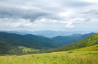 Dramatik ve bulutlu bir gökyüzünün altındaki dağ vadisi manzarası. Ön plan çimenli bir çayırdır, uzak dağlar ise mavi ve yeşil tonlarla kaplıdır. Karpatlar, Ukrayna