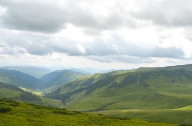 Parçalı bulutlu bir gökyüzünün altında geniş, çimenli dağ yamaçlarının panoramik manzarası. Yuvarlanan tepeler canlı bir yeşildir, manzarada küçük bir patika kıvrılır. Karpat Dağları, Ukrayna