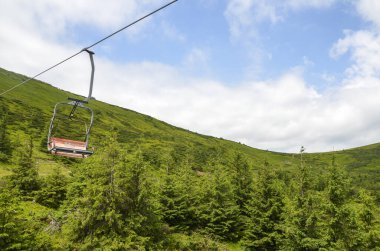 Gökyüzü bulutlu, yemyeşil bir tepenin altında, orman alanları ve doğal manzaralarla çevrelenmiş, açık hava macerası ve ulaşımın özünü yakalayan sandalye kaldırışının manzarası.