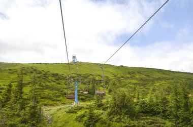 Gökyüzü bulutlu, yemyeşil bir tepenin altında, orman alanları ve doğal manzaralarla çevrelenmiş, açık hava macerası ve ulaşımın özünü yakalayan sandalye kaldırışının manzarası.