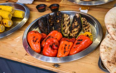 Ahşap bir masanın üzerindeki gümüş tabakta, kömürleşmiş patlıcan, kabak ve canlı kırmızı ve turuncu dolma biber içeren lezzetli bir çeşit ızgara sebze bulunur. 