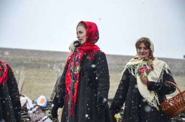 Parlak renkli başörtüleri ve kalın mantoları da dahil olmak üzere geleneksel Ukrayna kıyafetleri içindeki iki kadın Shrovetide kutlamaları sırasında dışarıda karlı bir ortamda dururlar.