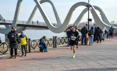 Erkek koşucu, benzersiz dalgalı bir köprünün yer aldığı manzaralı bir nehir kenarında yarışırken, seyirciler açık bir gökyüzü altındaki açık hava sahnesinin tadını çıkarıyorlar.
