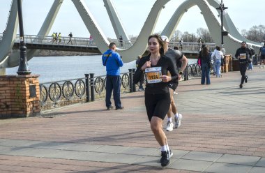 Açık ve güneşli bir günde nehir kenarındaki bir köprünün yakınındaki bir maraton yarışında koşan bir kadın açık hava sporları ve aktif yaşam tarzları sergiliyor.