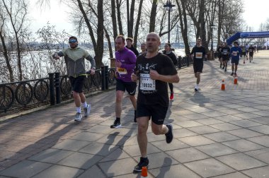 Dinamik görüntü, güneşli bir günde nehir kenarındaki patikada koşan bir grup koşucunun spor, açık hava aktivitesi ve toplumsal katılımı vurguluyor.