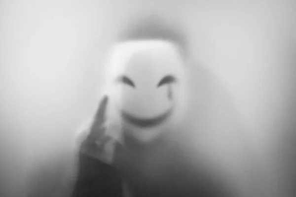 Bangkok, Thailand - August, 18, 2023 :Mysterious man in smiling mask stands behind frosted glass, spooky style, blurred image. Halloween concept.