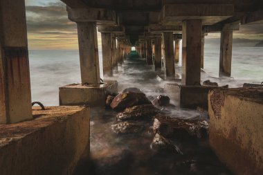 Kayalık bir sahil manzarası dramatik bir gün batımında eski bir rıhtıma yanaşıyor..