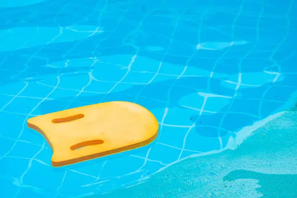 Yellow kick board made of foam floating on water surface in swimming pool.