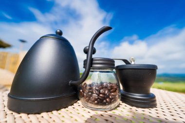 This captivating image captures the essence of a serene morning ritual. On an outdoor table, a sleek black teapot and a matching manual coffee grinder with a silver handle are prominently displayed. A glass jar filled with whole coffee beans sits bes clipart