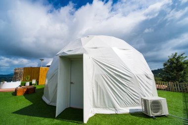 Modern bir jeodezik kubbe, yemyeşil bir çimenlikte, geniş mavi bir gökyüzünün altında, tüylü beyaz bulutlarla noktalanmış.