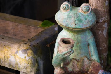 Bir ağaç ve yıpranmış bir bankın yanına yerleştirilmiş, elinde fincan tutan büyüleyici bir kurbağa dekoru. Dekorun yaşlı görünümü karakter katıyor..