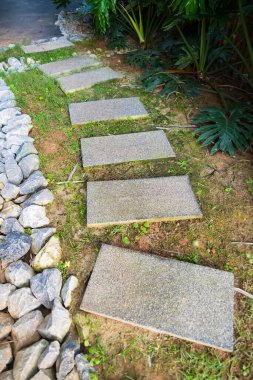 A serene garden pathway made of rectangular stone slabs, surrounded by lush greenery and decorative rocks. clipart