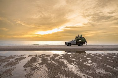Bir plajda çöp toplama kamyonu ve iki temizlik işçisi çöp konteynerinin üstünde otururken görülüyor. Sahne, plajdaki atıkları yönetme ve temizliği koruma çabalarını vurguluyor.