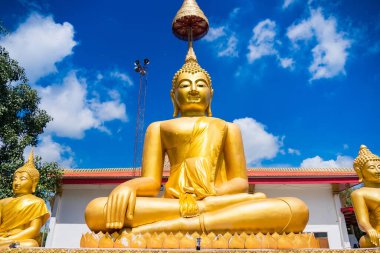 Huzurlu, altın bir Buda heykeli. Meditasyon pozu veren bir heykel. Canlı, mavi bir gökyüzünün altında, beyaz bulutlu..