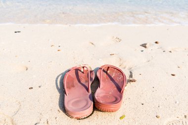 Deniz kenarındaki huzurlu anları sembolize eden kırmızı parmak arası terliklerin olduğu sakin bir sahil sahnesi..